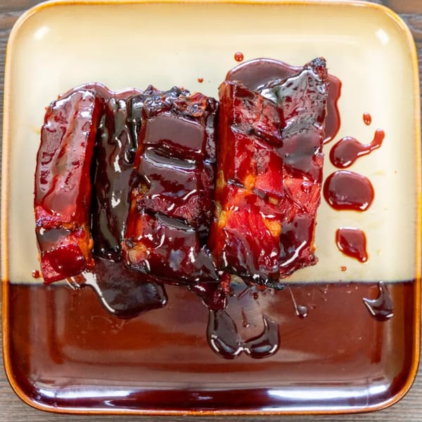 Four pieces of dark red, glazed BBQ ribs on a two-toned brown and tan plate.