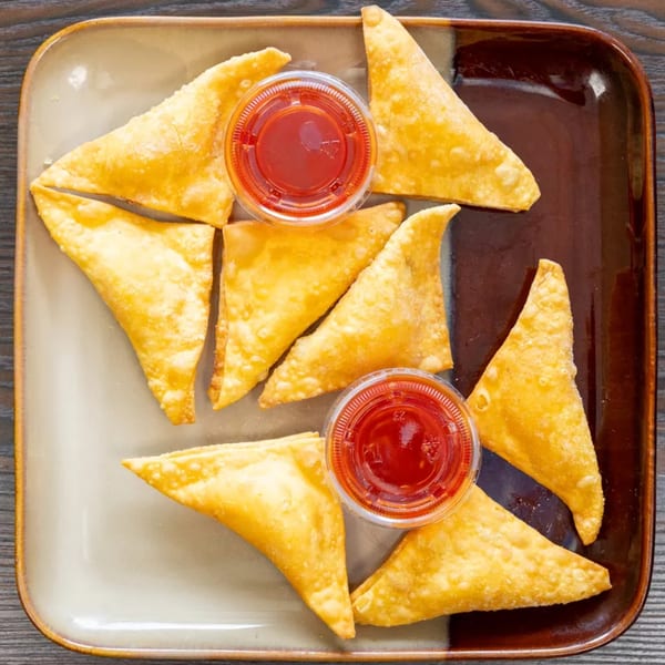 Golden fried wontons arranged on a two-toned rectangular plate with two small cups of red dipping sauce.