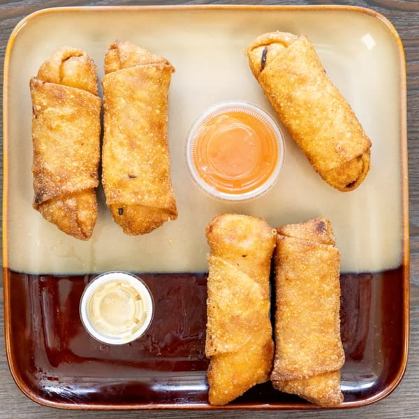 Five crispy golden-brown egg rolls arranged on a two-toned brown and tan rectangular plate with two dipping sauces.